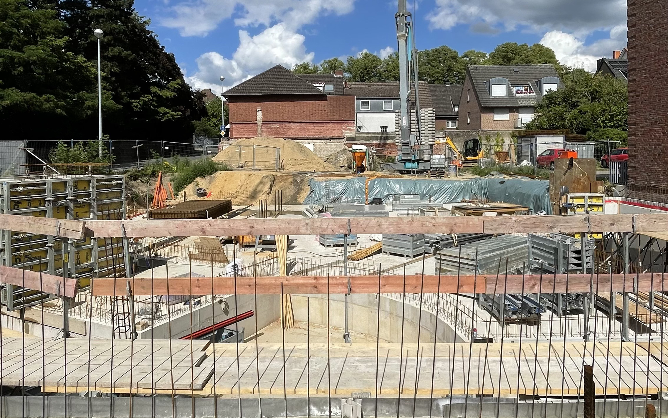 Baustelle mit frisch betonierter Bodenplatte, Baustellenmaterialien und Schalungselementen. Im Hintergrund stehen Maschinen, Baumaterialien und Wohnhäuser. Das Baufeld ist eng und von umliegender Bebauung eingefasst.